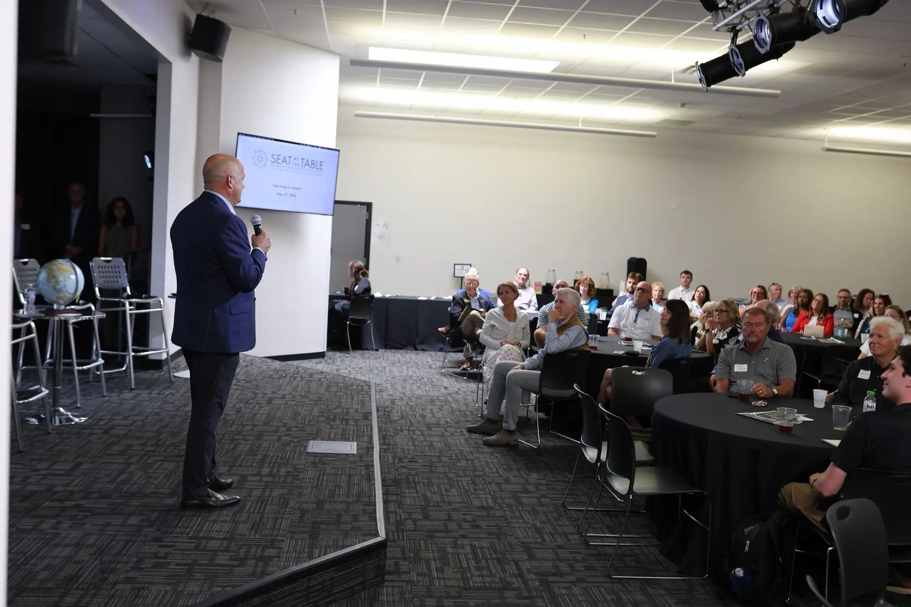A speaker addressing the full room from the front, guests at round tables